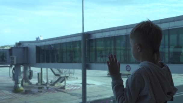 Kleine jongen op zoek naar venster terwijl het wachten vliegtuig in de vertreklounge in luchthaven. Jongen op zoek naar vliegtuig van de luchthaven terminal-venster in de wachtruimte. Vakantie en vakantie concept. — Stockvideo