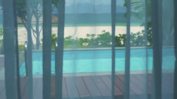 View from window with curtains on swimming pool and sea beach. Luxury villa with swimming pool, blue sea and beach landscape with palm tree. — Stock Video