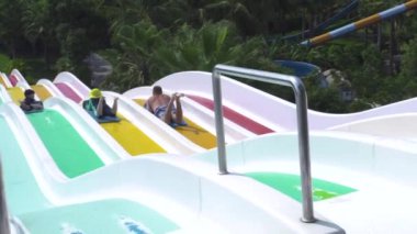 İnsanlar yaz tatilinde eğlence aquapark renkli su kaydırağı sürme. Yeşil palmiye ağacı manzara açık su parkında slaytlar üzerinde kayan eğlenen insanlar.