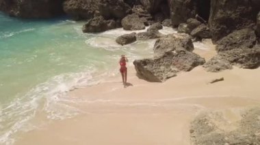 Bikinili genç bir kadın kumlu sahilde deniz dalgaları ve kayalık kayalıklarda yürüyor. Tropikal plajdaki İHA 'lı çekici kadından hava görüntüsü. Mavi deniz dalgaları taşlı kıyılara vuruyor.