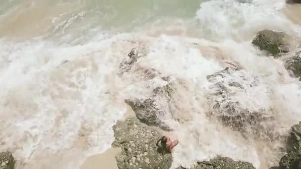 Young woman in bikini on sea shore and splashing water waves aerial landscape. Sea waves splashing with foam and spray to stony coast. Beautiful woman on rocky sea beach drone view. — 비디오