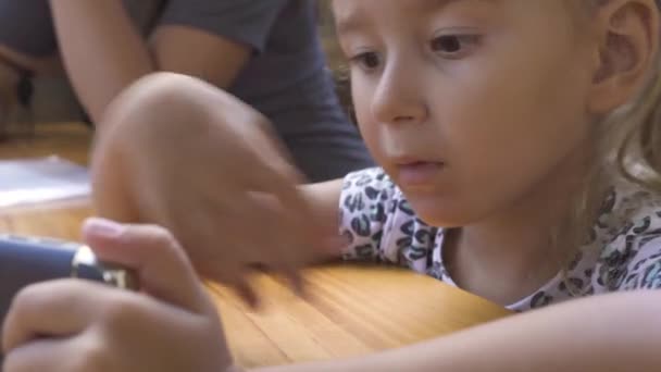 Retrato linda niña buscando teléfono móvil sentado en la mesa. Cerca de la cara divertida chica usando el teléfono inteligente para ver películas de dibujos animados . — Vídeo de stock