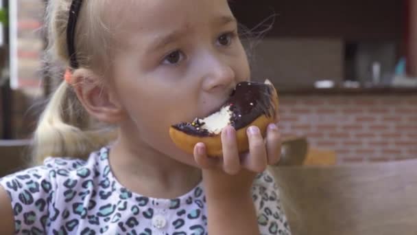 Забавна дівчинка їсть солодкий шоколадний пончик за столом у кафе. Обличчя мила блондинка кусає шоколадний пончик в десерті в ресторані . — стокове відео