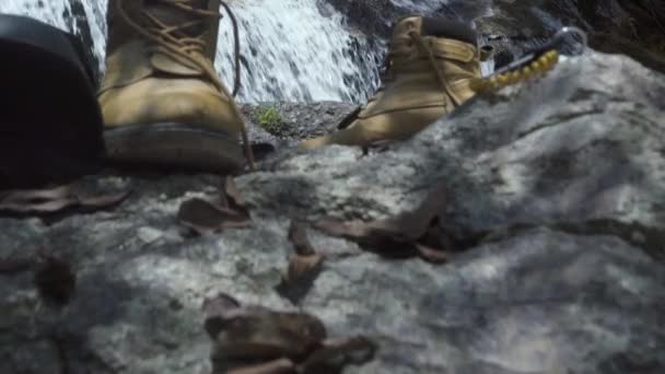 山の流れる滝の上に石の上に立つ男性の靴。山の滝で入浴する観光客。熱帯雨林で新鮮な山の川を楽しむハンサムな男. — ストック動画