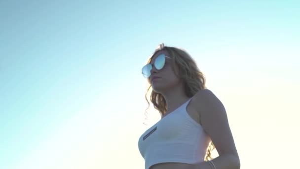 Σέξι ξανθιά κοπέλα που φοράει μπλούζα καλλιέργειας, γυαλιά ηλίου, θέα κάτω γωνία. φόντο ουρανού — Αρχείο Βίντεο