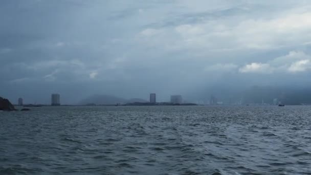 Vue du milieu de la mer ondulée, nuages sombres sur une ville et des montagnes brumeuses. — Video