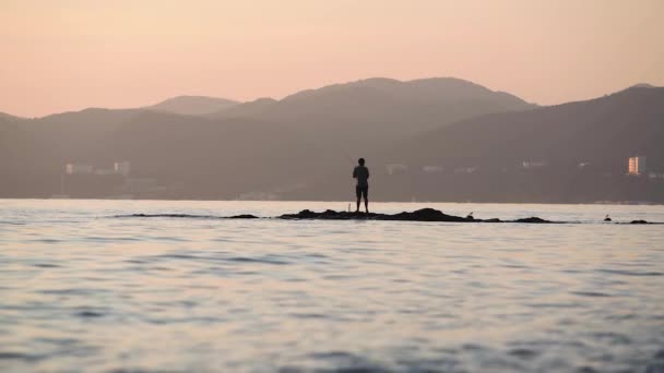 Unregconizable hombre de pie y la pesca en las rocas en medio del mar. — Vídeo de stock