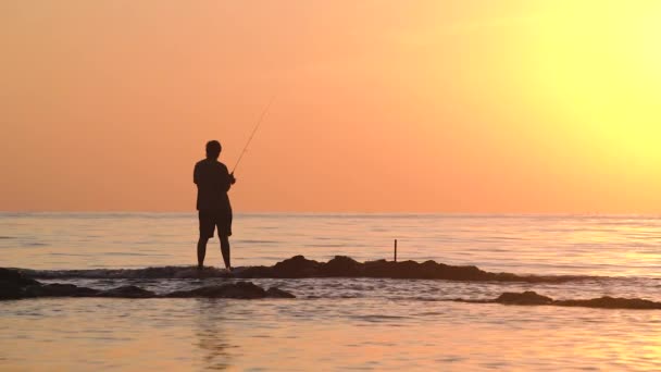 Silueta de un hombre de pie sobre las rocas en el mar sosteniendo una caña de pescar. — Vídeos de Stock