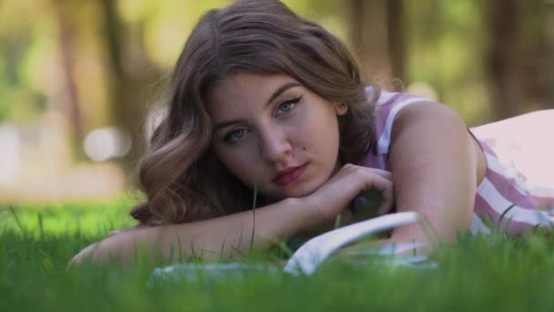 Ung kvinna med ljust hår ligger på grönt gräs nära bok — Stockvideo
