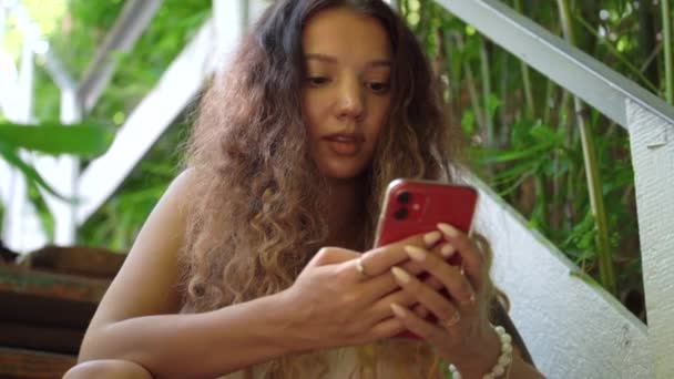Elegante mujer joven con el pelo rizado envía SMS en el teléfono inteligente — Vídeos de Stock