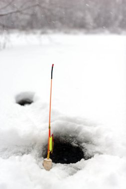 Homemade winter fishing rod stands at the hole in the ice of the river against the snow-covered forest. clipart