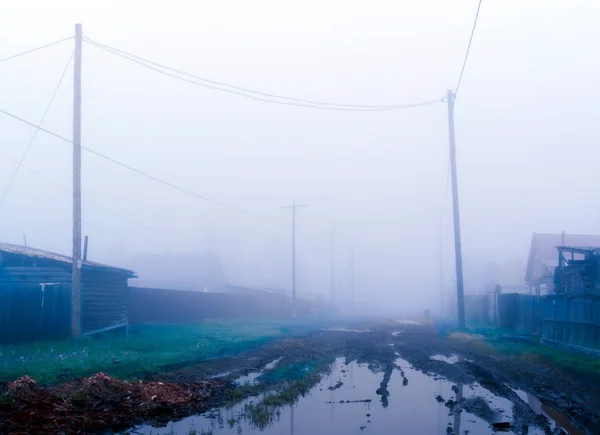 A large puddle on a village street in the morning heavy fog among the houses with fences and poles with power wires in the North of Yakutia.