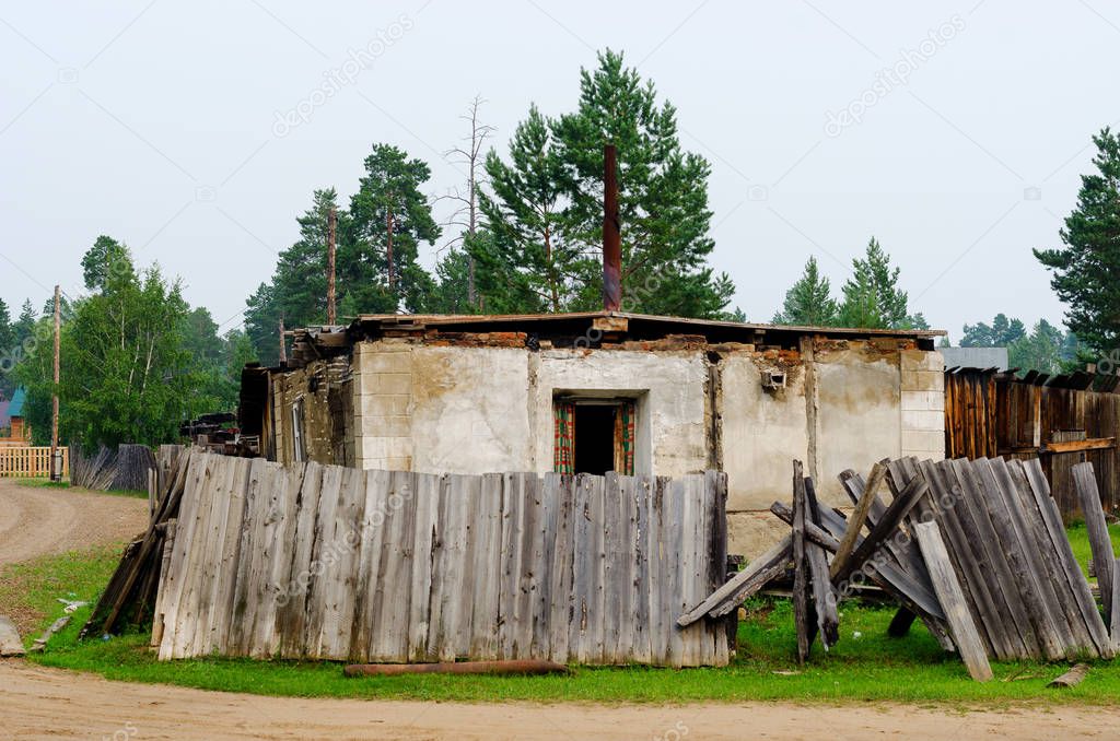An old, rickety wooden barn stands with an open door behind a fallen fence in the Northern village of Yakutia against a forest of pines.