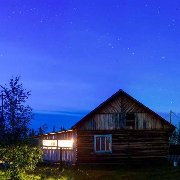 The bright light of the lamp from the veranda of a private house illuminates the birch and grass standing at night under the sky with stars in the wild North of Yakutia