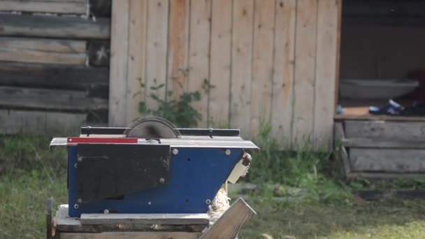 De aandrijving van de Planer en een cirkelzaag spinnen zonder planken met zaagsel vliegen in de achtergrond van de vervaagde muren van houten huizen en gras. — Stockvideo