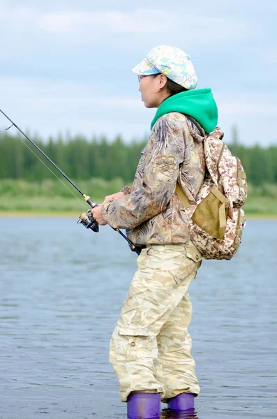 雅库特女孩旅游渔夫看着钓鱼竿钓鱼在岸上旋转与背包在河和冻土云杉森林的帽子和靴子 — 图库照片
