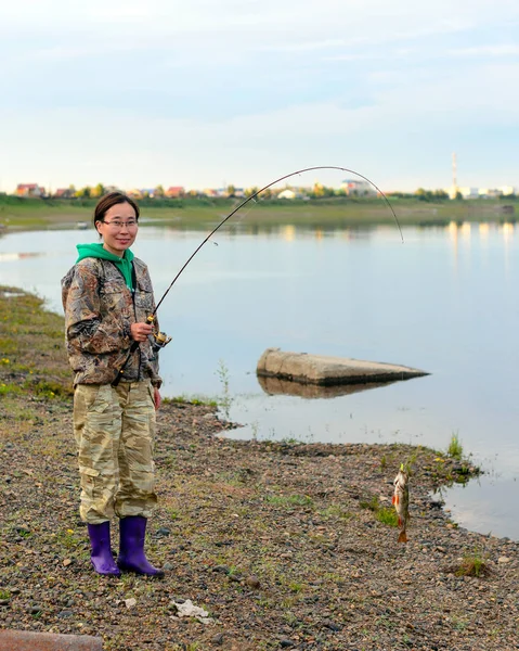 ヤクートの女の子観光漁師は 川のほとりに立って 曲がった回転棒の上にゴムのルアーにぶら下がっている魚のパーチのキャッチと村の家 — ストック写真