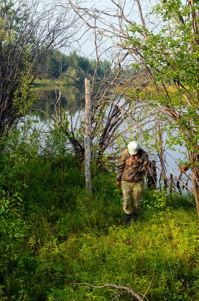 保護色の服を着たヤクートの野生の森の枝から顔を隠して湖から離れた少女観光客 — ストック写真