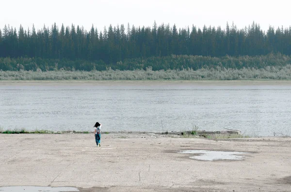 Girl traveler with backpack goes to the edge of the concrete sidewalk touching the hair on the Bank of the old pier North of the river vilyu with spruce forests and the tundra of Yakutia on the other side.