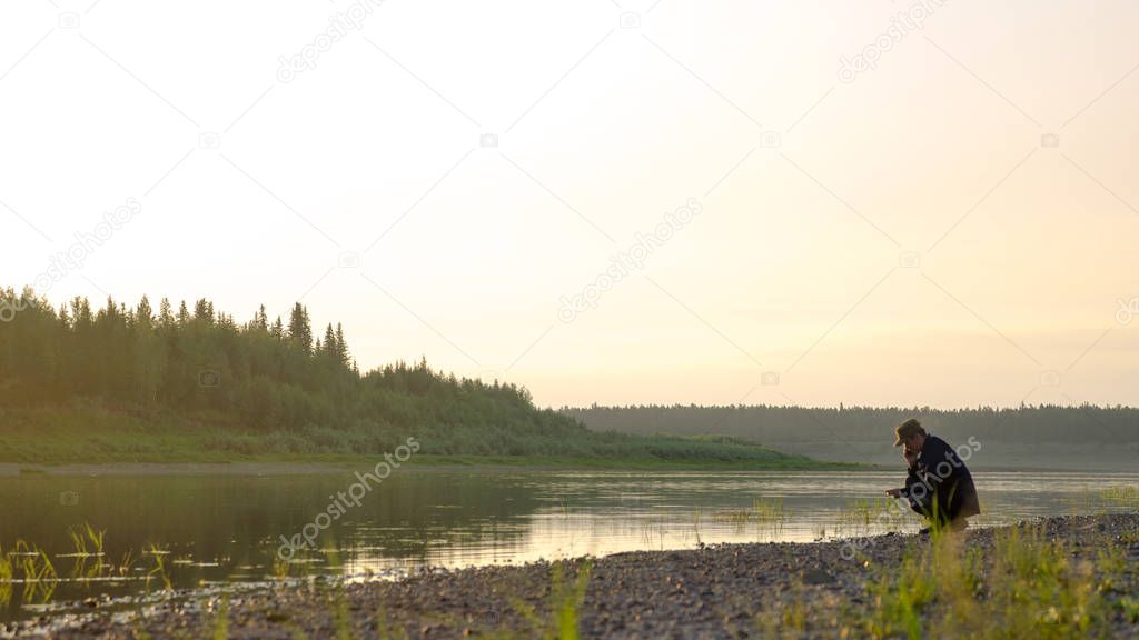 The young man Yakut squatting sklona head talking on the mobile phone on the shore of the wild North of the Vilyui river in the forest.