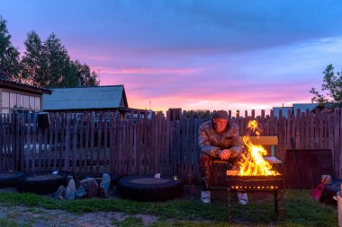 An old lonely man sits sadly on a bench at a village house behind a fence looking into the fire of a barbecue fire at sunset in the evening. clipart