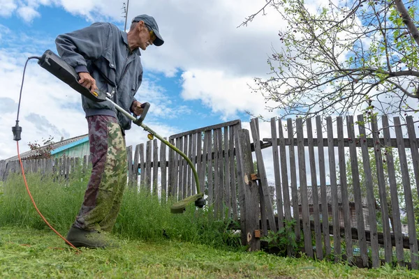 An old man with an electric lawn mower trimmer cuts fresh green grass by the fence wearing glasses and a cap in the village.