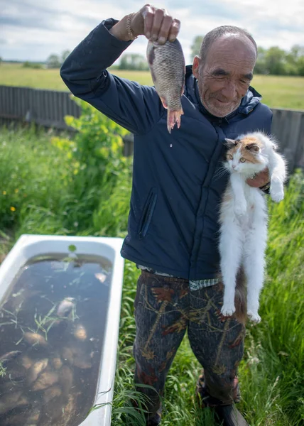 A village old man holds a carp in one hand and a cat looking at food in the other against the background of a bath with fish behind the fence.