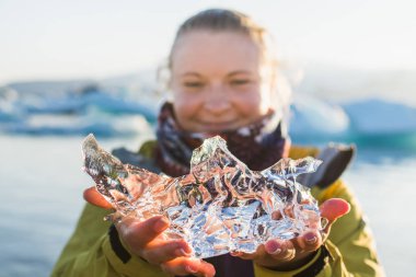 Yüzünün önünde şeffaf bir parça Jokulsarlon gölünde, İzlanda tutan genç kadın. Bulanık arka plan.