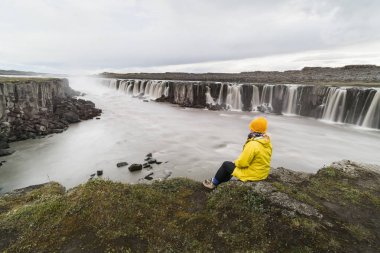 Kadın sarı yağmurluk oturma ayakta Vatnayokull Milli Parkı, İzlanda Selfoss şelale yanındaki uçurumun kenarında.
