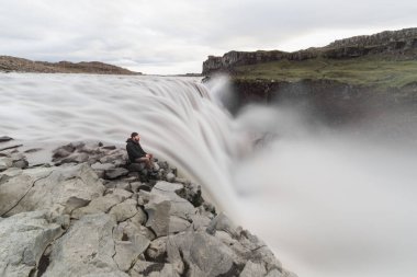 Adam oturma ayakta Vatnayokull Milli Parkı, İzlanda Dettifoss şelale yanındaki uçurumun kenarında. Bulutlu gün, uzun pozlama.
