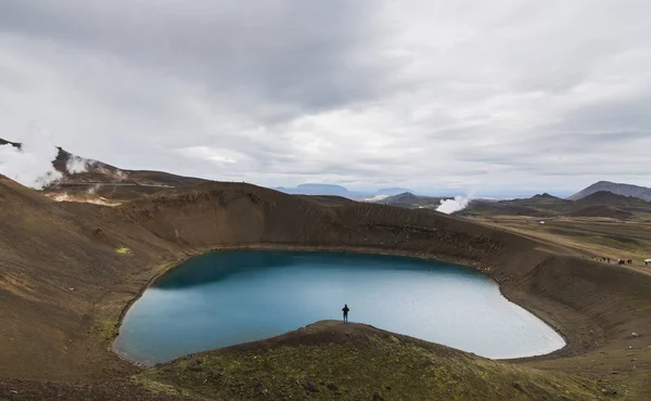 Human figure standing in the water filled crater of Krafla volcano, Iceland. Panoramic shot, cloudy day.