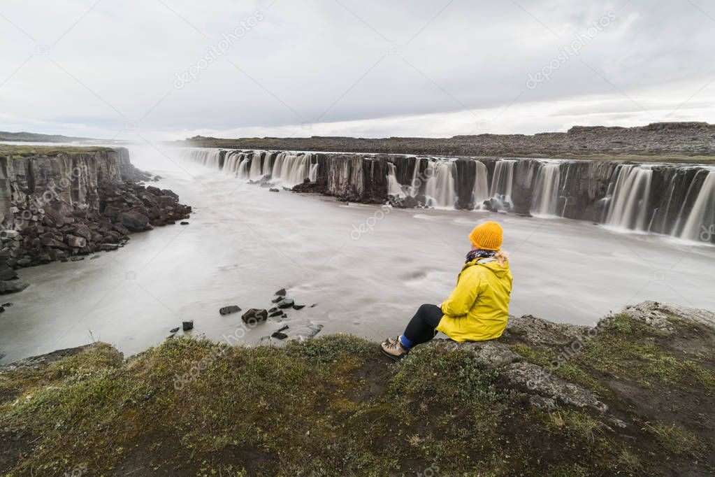 Woman in yellow raincoat sitting standing on the cliff edge next to Selfoss waterfall in Vatnayokull national park, Iceland.