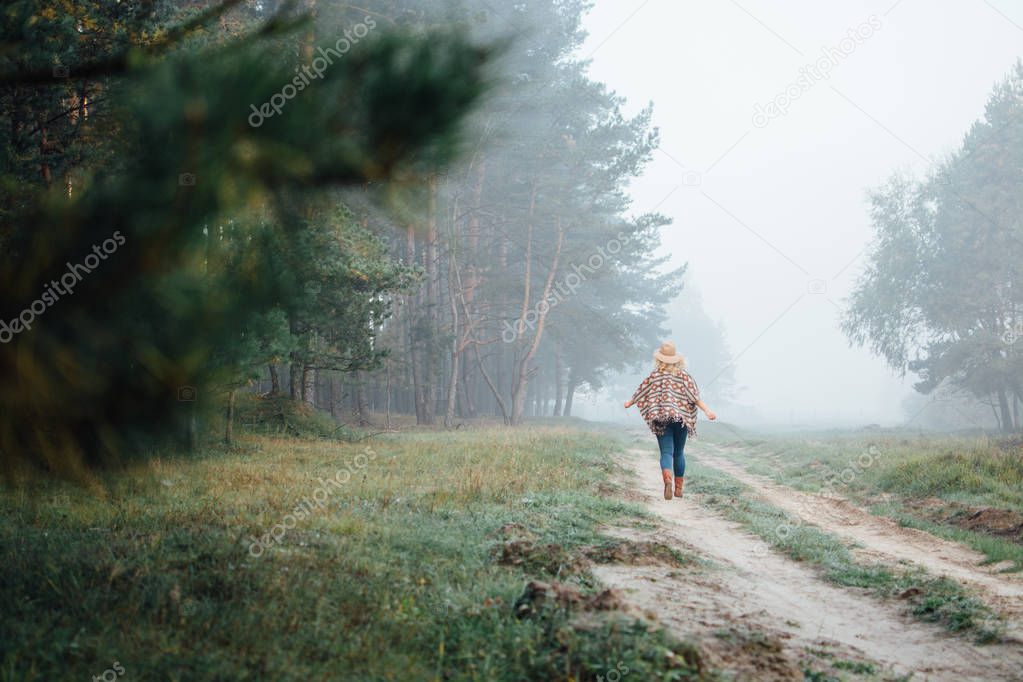 Woman in wide-brimmed felt hat and authentic poncho going along the forest at foggy morning.