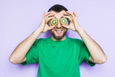 Genç gülümseyen ve sürpriz onun gözlerinin önünde yeşil kivi dilimlerini tutan adam sakallı. Işık arka plan mor, alanı Kopyala.