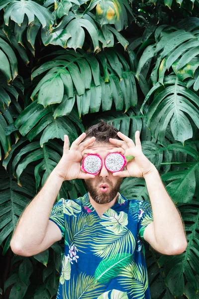 Jonge man met baard die plakjes Pitaya drakenfruit voor zijn ogen houdt, verrast. — Stockfoto