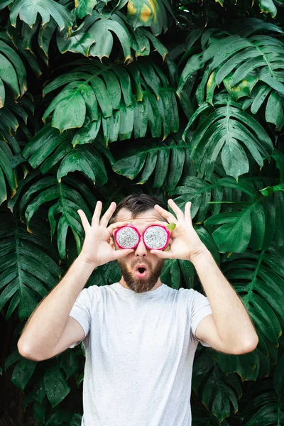 Jonge man met baard die plakjes Pitaya drakenfruit voor zijn ogen houdt, verrast. — Stockfoto