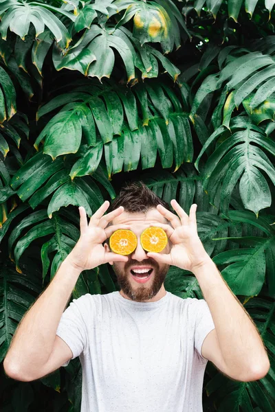 Jonge man met een baard die stukjes oranje mandarijn voor zijn ogen houdt, lachend — Stockfoto