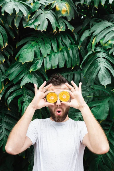 Een jonge man met een baard die stukjes oranje mandarijn voor zijn ogen houdt, verrast. — Stockfoto