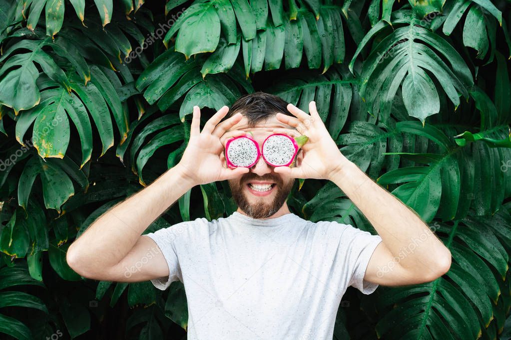 Young bearded man holding slices of Pitaya dragon fruit in front of his eyes, laughing