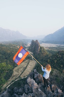 Sarışın kadın Vang Vieng, Laos vadi bakan elinde Laos bayrağı ile dağ zirvesinde duruyor