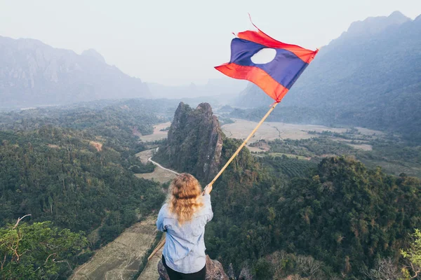 ブロンドの女性は、ラオスのヴァンヴィエンの谷を見下ろす彼女の手の中にラオスの旗を持つ山の頂上に立っています — ストック写真