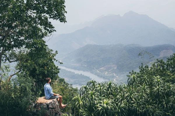 Joven hombre caucásico sentado en la cima de la montaña con vistas al valle del río en el pueblo de Nong Khiaw, Laos — Foto de Stock