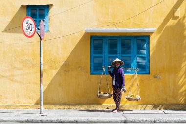 Hoi An, Vietnam - Haziran 2019: sokak meyve satıcısı kadın eski şehirde renkli evin yanından yürüyor
