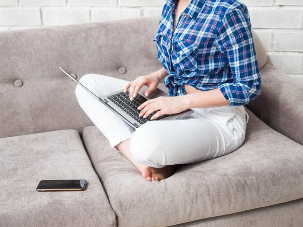 A mulher está a trabalhar sentada no sofá. As mãos estão segurando o telefone celular e trabalhando com um notebook . — Fotografia de Stock