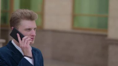 Yakışıklı genç iş adamı bir klasik takım elbise ve ceket iş bölgesi sokaklarında yürüyüş akıllı telefon üzerinde konuşuyor