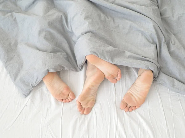 Feet of couple sleeping side by side in comfortable bed. Close up of feet in a bed under white blanket. Bare feet of a man and a woman peeking out from under the cove.