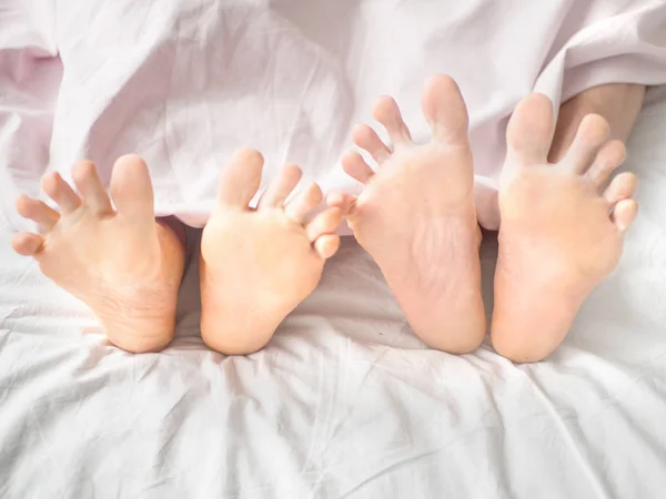 Stäng upp barfota ben av ett par på vit säng i sovrummet på morgonen — Stockfoto
