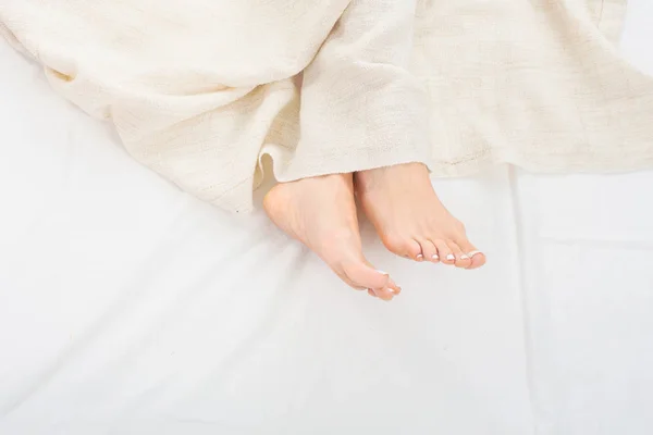 Female feet under blanket flat lay. Female beautiful feet with red pedicure on the bed. Top view on the sleeping woman legs under blanket