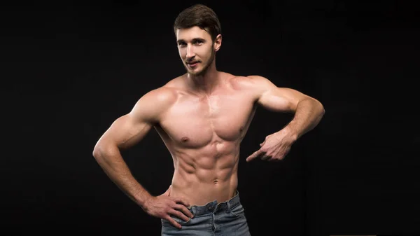 Guapo joven musculoso hombre sin camisa con jeans, sobre fondo oscuro en la toma de estudio. Mostrando su ABS con el dedo — Foto de Stock