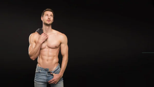 Un bel giovane uomo forte sessuale con corpo muscoloso in jeans blu con camicia sulla spalla in posa in studio su sfondo scuro — Foto Stock